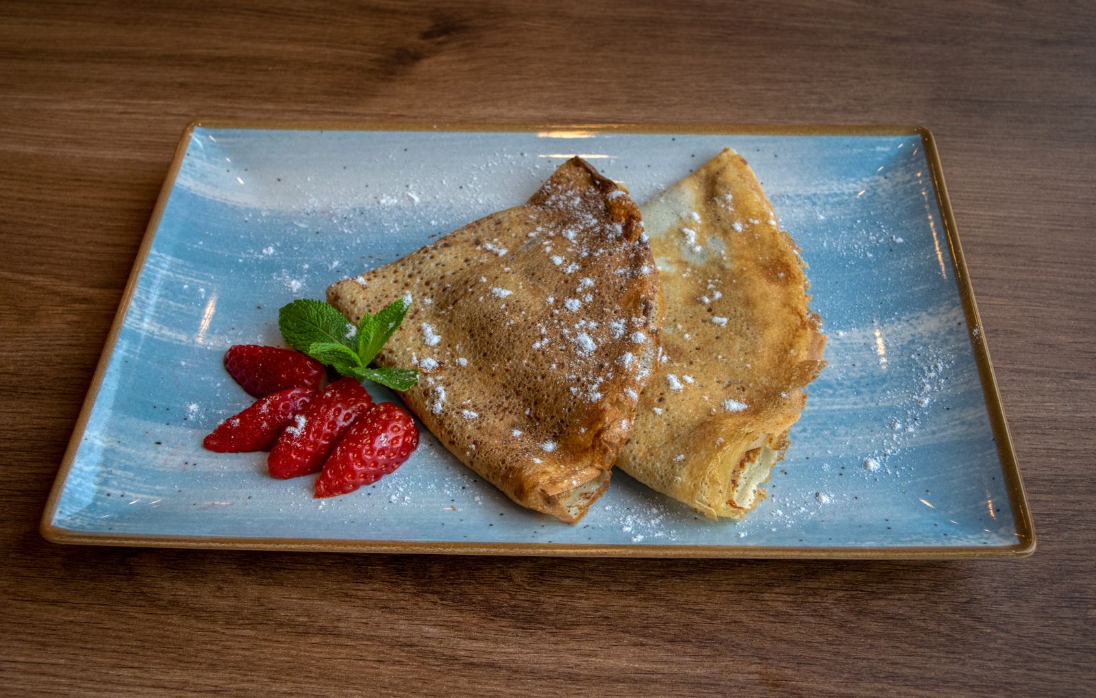 Zwei gefaltete Crêpes mit Erdbeeren und Puderzucker auf blauem Teller